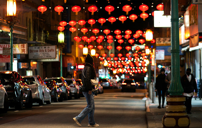 A pedestrian walking down a street in San Francisco's Chinatown illuminated by rows of glowing red lanterns at night