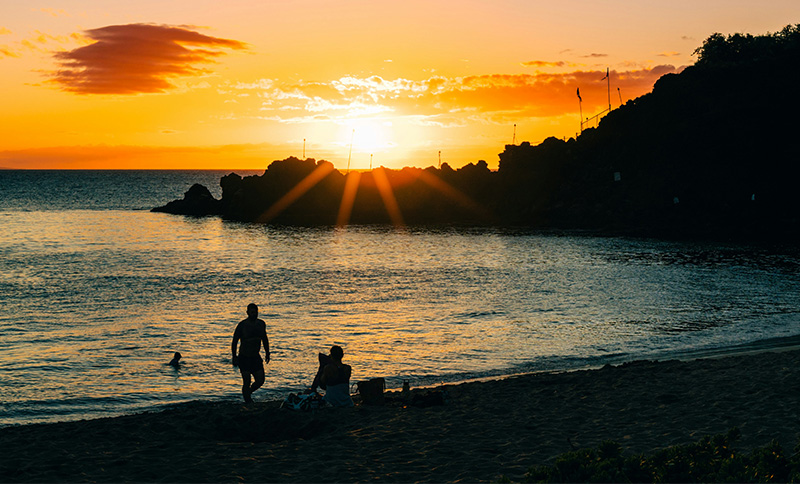 Silhouettes of people relaxing on the sand and wading in the water at a quiet beach during a vibrant, golden sunset.