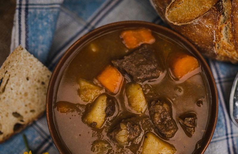 A bowl of rich, dark Irish beef stew filled with tender chunks of meat, large slices of carrots, and a whole potato, served in a rustic black ceramic bowl on a wooden tabletop