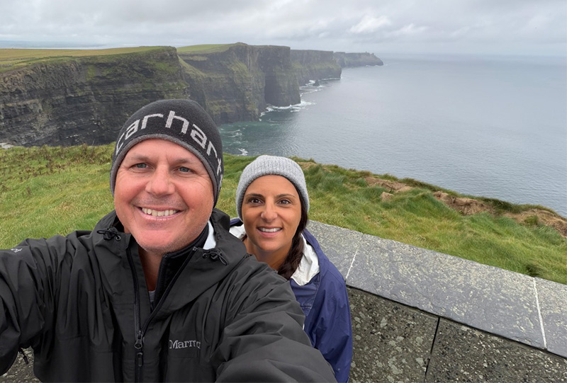 A smiling man and woman taking a selfie in front of the scenic Cliffs of Moher in Ireland on a cloudy day.