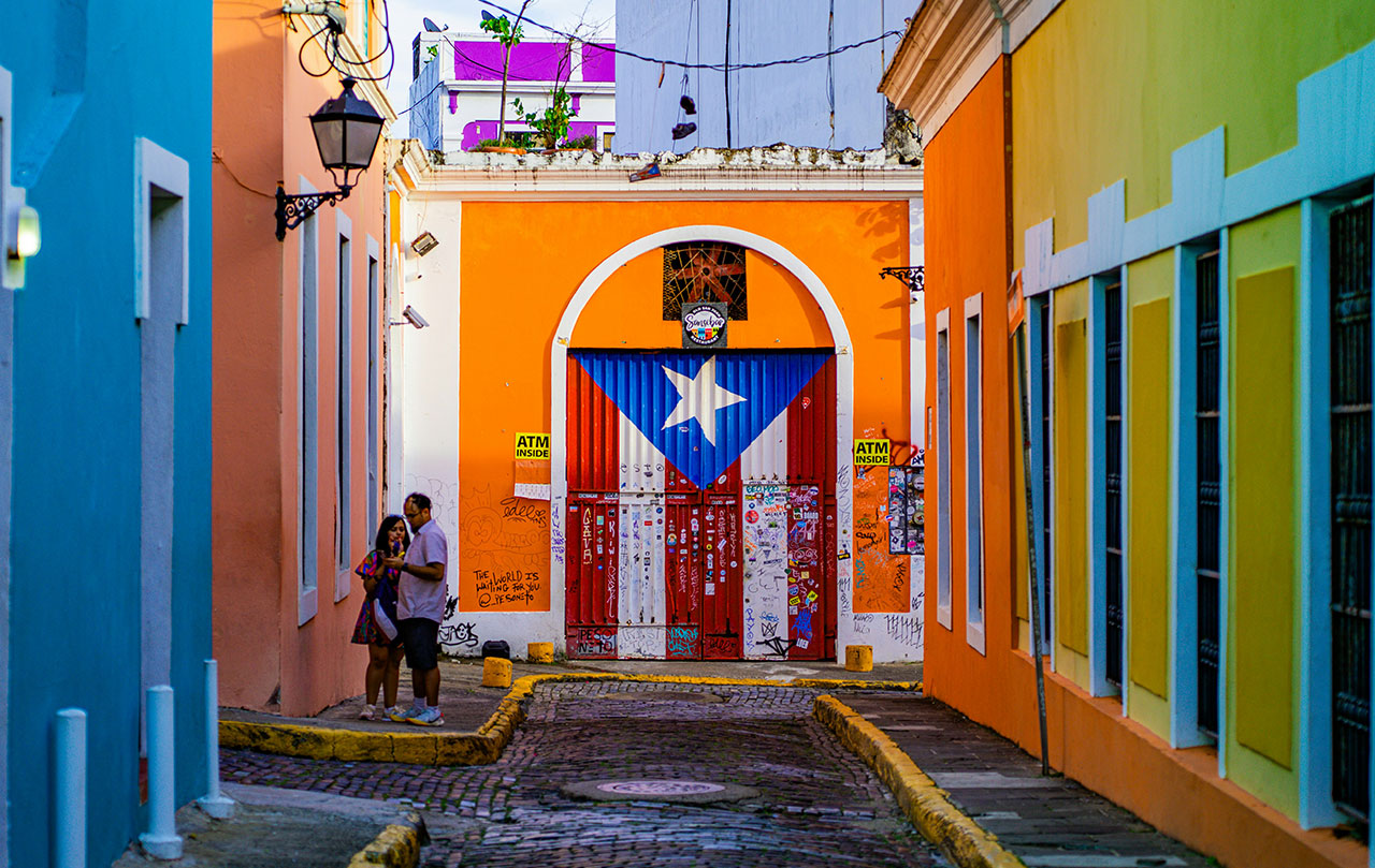 A narrow cobblestone street in Old San Juan, Puerto Rico, lined with vibrant orange and yellow colonial buildings featuring blue doors and balconies, with a large Puerto Rican flag painted on a central door.