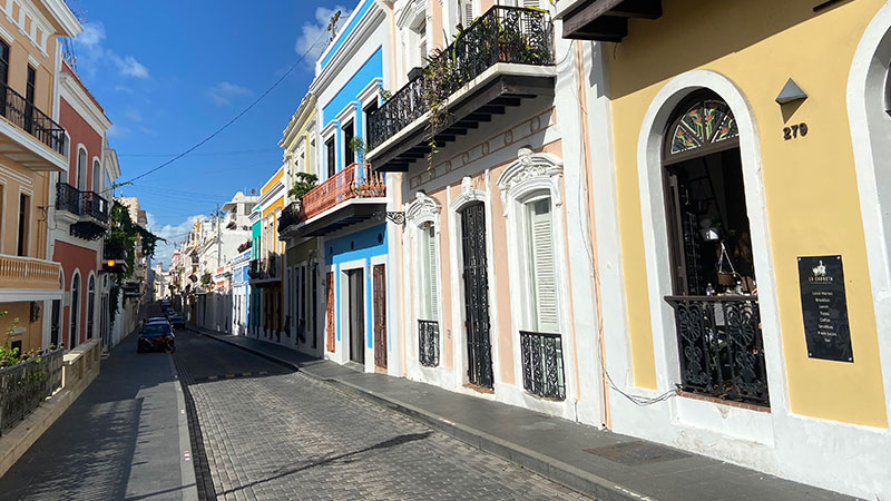 A bright, sunlit cobblestone street in Old San Juan lined with colorful colonial buildings and balconies.