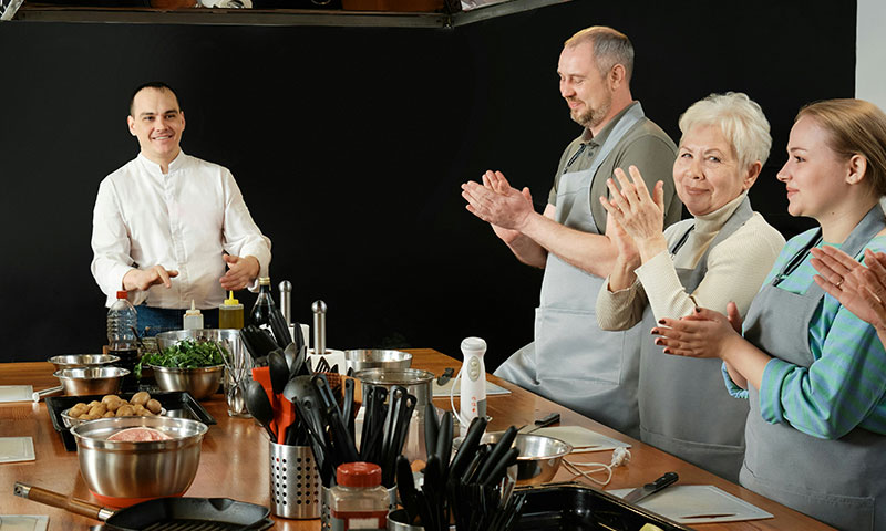 A chef in a white uniform stands behind a wooden kitchen island while three students in gray aprons applaud during a cooking class.