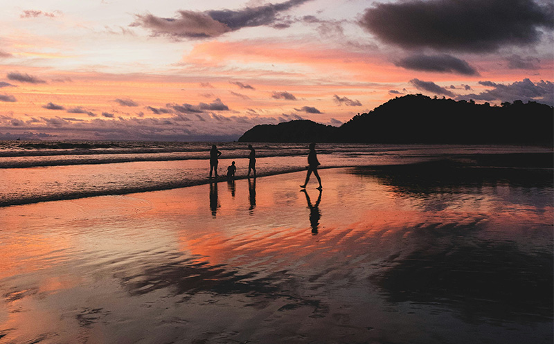 An expat couple walking along a tropical beach at sunset, with the orange and purple sky reflecting on the calm water and wet sand.