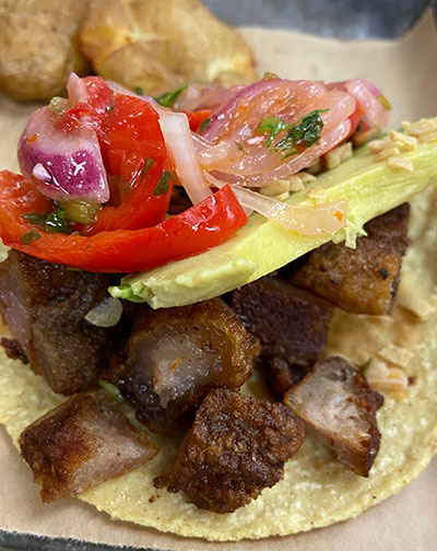Close up of a street taco with crispy chunks of fried pork, a fresh slice of avocado, and vibrant red salsa on a corn tortilla.