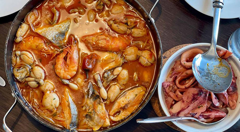 A large shallow pan filled with traditional Croatian seafood stew featuring shrimp, fish, and mussels in a tomato broth, served next to a bowl of cooked squid.