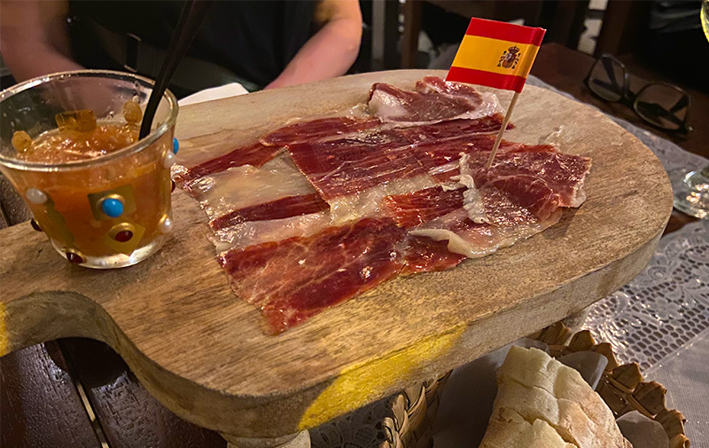Thinly sliced Serrano ham served on a raised wooden board with a miniature Spanish flag, fresh bread, and tomato puree at Doña Lola in Cartagena