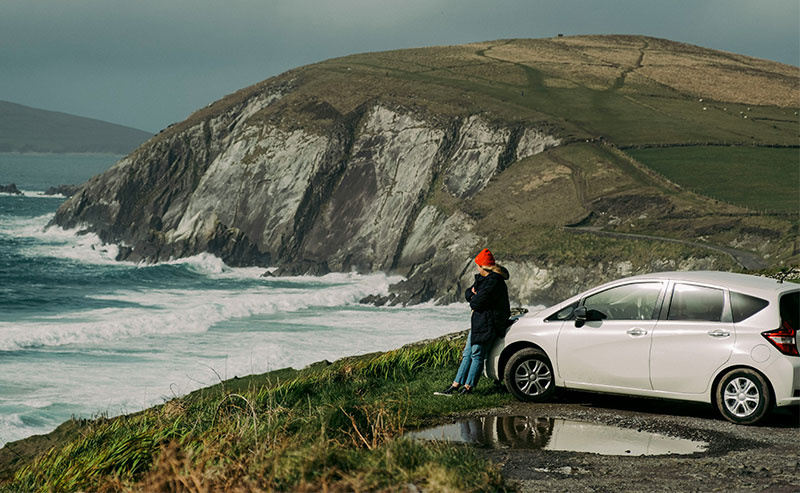 A view from the driver’s seat of a car traveling along a winding, narrow coastal road toward Dingle in County Kerry, Ireland, with lush green hills on one side and the deep blue Atlantic Ocean on the other.