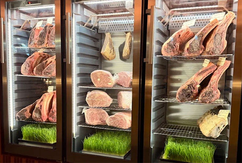 A professional display of various cuts of dry-aged beef arranged on metal racks inside a glass-fronted aging room at Bazaar Meat by José Andrés