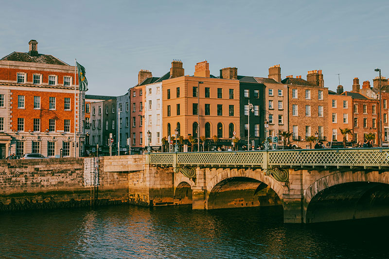 A view of the Ha'penny Bridge at dusk with its white iron arches illuminated, reflecting on the surface of the River Liffey as the city lights of Dublin begin to glow in the background.