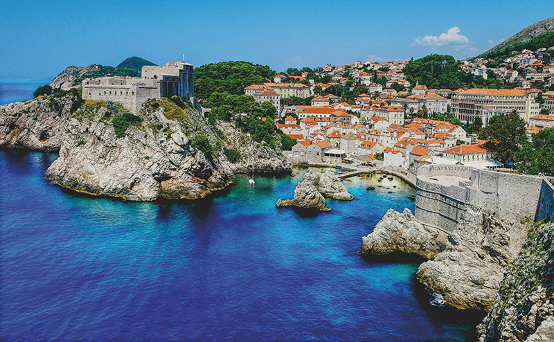 View of the rocky Dubrovnik West Harbor with clear blue water, Lovrijenac fortress on a cliff, and the walled city in the background.