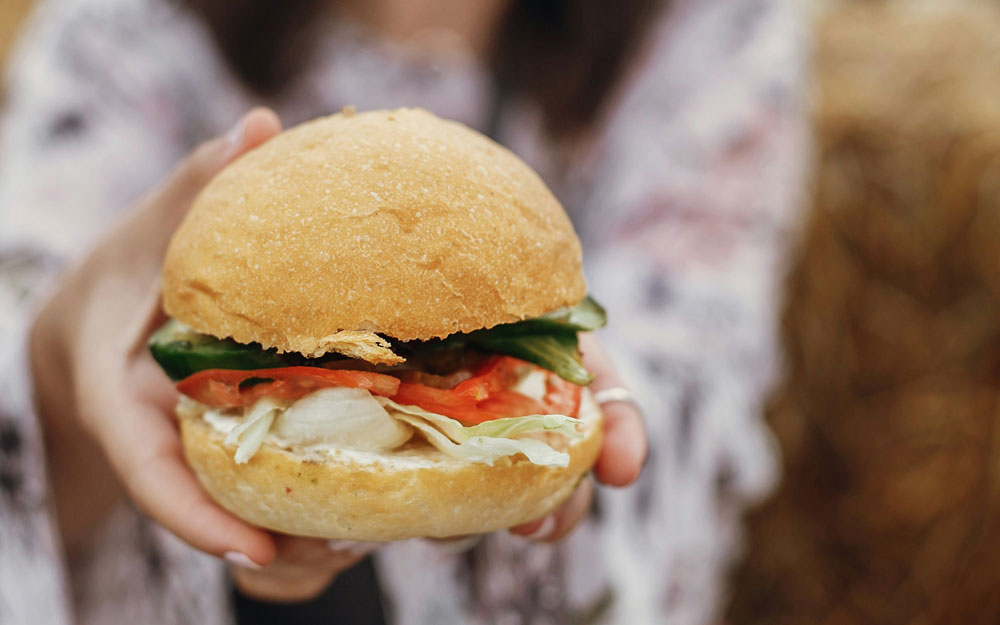 A hand holding a freshly made deli sandwich with turkey, sprouts, and tomato on a rustic roll, with a blurred outdoor background.