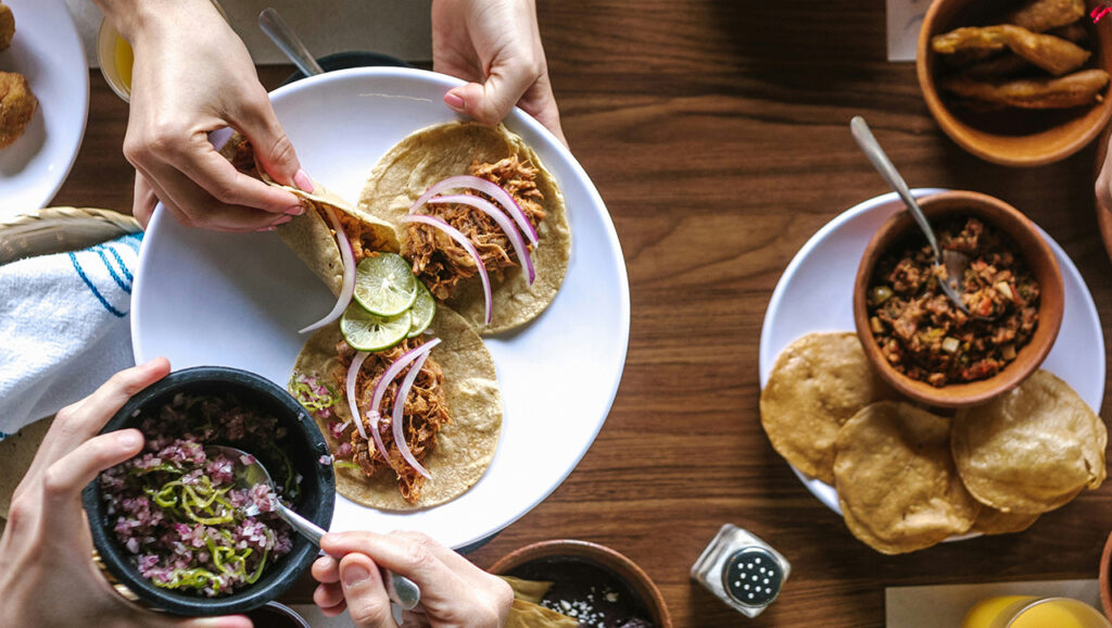 A top-down view of a rustic wooden table featuring several small plates of authentic local cuisine, including street tacos with onions and cilantro, small bowls of sauces, and a person's hands reaching for a dish.