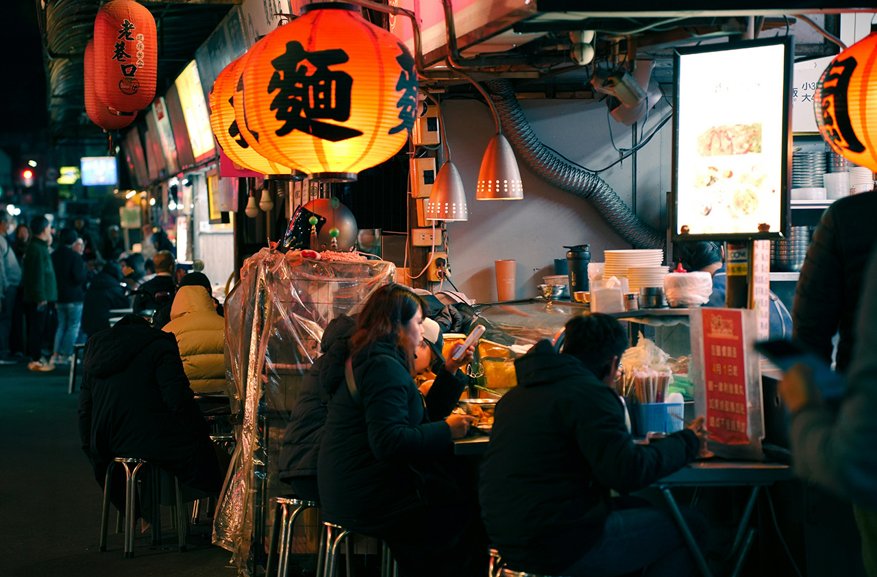 A bustling night market scene in Taipei, Taiwan, where locals and travelers are seated at small outdoor tables under glowing red and yellow lanterns, enjoying street food in a vibrant, atmospheric setting.
