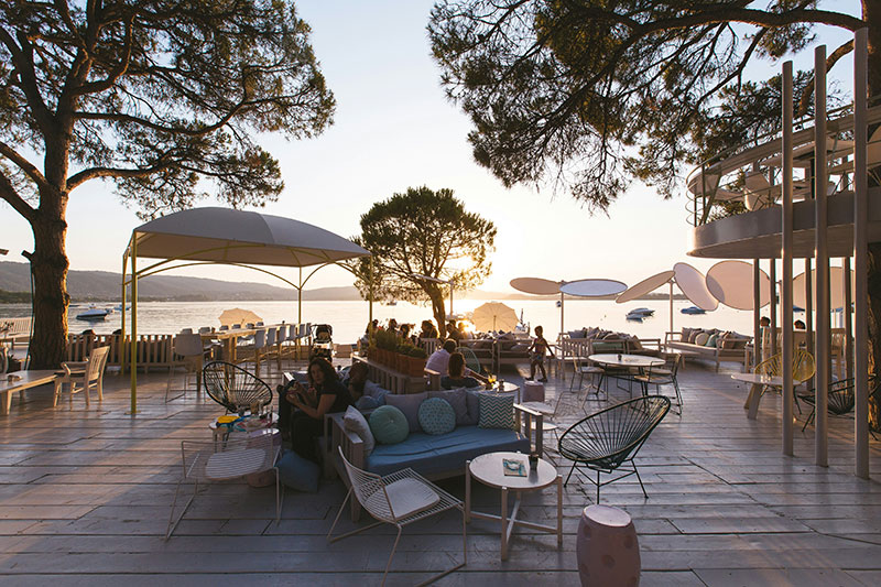 A serene, modern outdoor lounge area at Ekies All Senses Resort in Vourvourou, featuring minimalist white furniture, soft textiles, and a lush green backdrop of pine trees and Mediterranean plants.