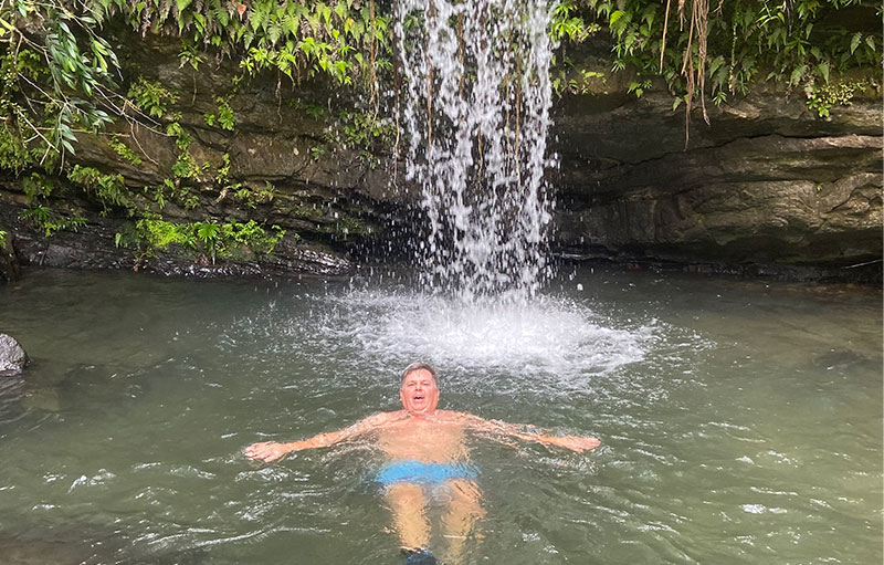 A man floating happily in a clear natural pool at the base of a beautiful rushing waterfall in the rainforest.