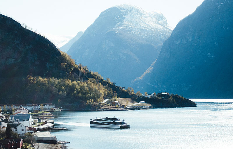 A modern white sightseeing ferry cruising through a deep blue fjord surrounded by steep, sunlit mountains in Norway.