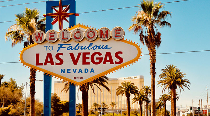 he iconic "Welcome to Fabulous Las Vegas" neon sign stands against a clear blue sky, surrounded by palm trees and desert landscaping.