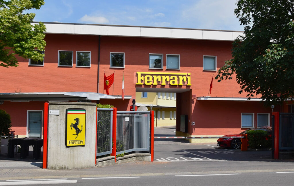 The historic red brick entrance of the Ferrari factory in Maranello, Italy, featuring the iconic yellow Ferrari shield and "Ferrari" signage.