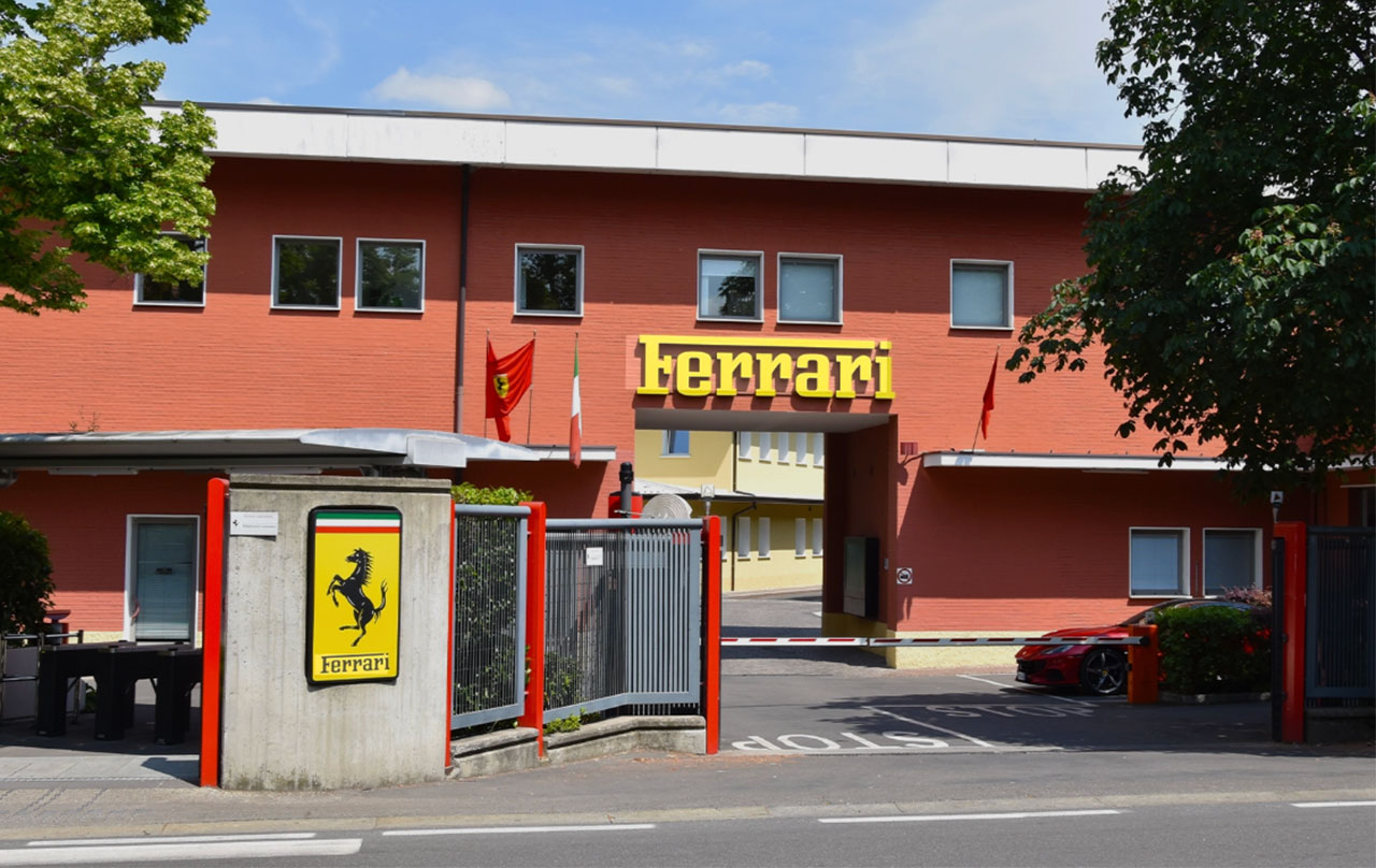 The historic red brick entrance of the Ferrari factory in Maranello, Italy, featuring the iconic yellow Ferrari shield and "Ferrari" signage.