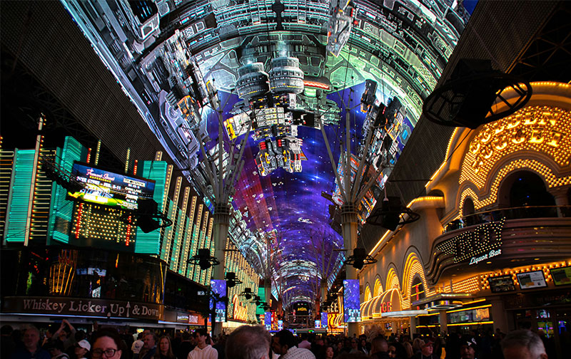 A bustling crowd walks beneath the massive, brightly lit LED canopy of the Fremont Street Experience in downtown Las Vegas at night.