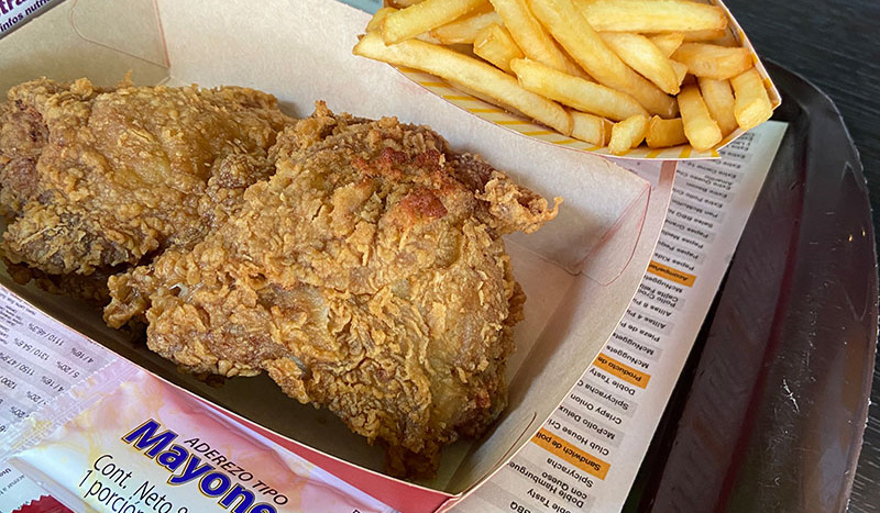 A close-up of a classic Frisby fried chicken meal in Cartagena, featuring a large piece of golden-brown breaded chicken served with a side of French fries in a branded paper container.