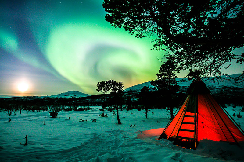 A small, glowing orange tent sits on a snowy plain in Abisko, Sweden, under a vibrant green Aurora Borealis dancing across the starry night sky.