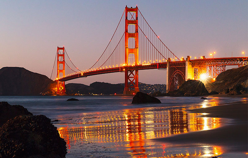 The illuminated Golden Gate Bridge glowing red against a twilight sky viewed from a rocky beach.