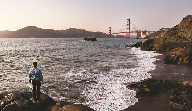 A person standing on a rocky shoreline looking out across the water at the Golden Gate Bridge during a warm sunset