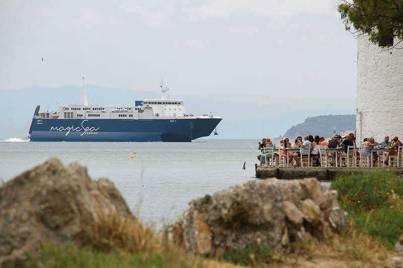 A view from a rocky coastline shows a large blue and white passenger ferry, named "SEA JETS," steaming across the blue water towards an island port. In the foreground, dry grasses and scrub cover the rocks.