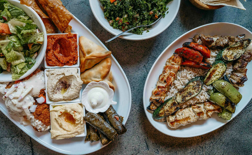 top-down spread of traditional Greek dishes including a Greek salad with a large block of feta, stuffed grape leaves (dolmades), grilled meats, pita bread, and various dips like tzatziki and hummus.