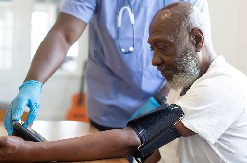 A medical professional in blue scrubs and gloves wraps a blood pressure cuff around the arm of an elderly man during a checkup. The scene is bright and professional, emphasizing high-quality healthcare and routine medical monitoring for seniors.