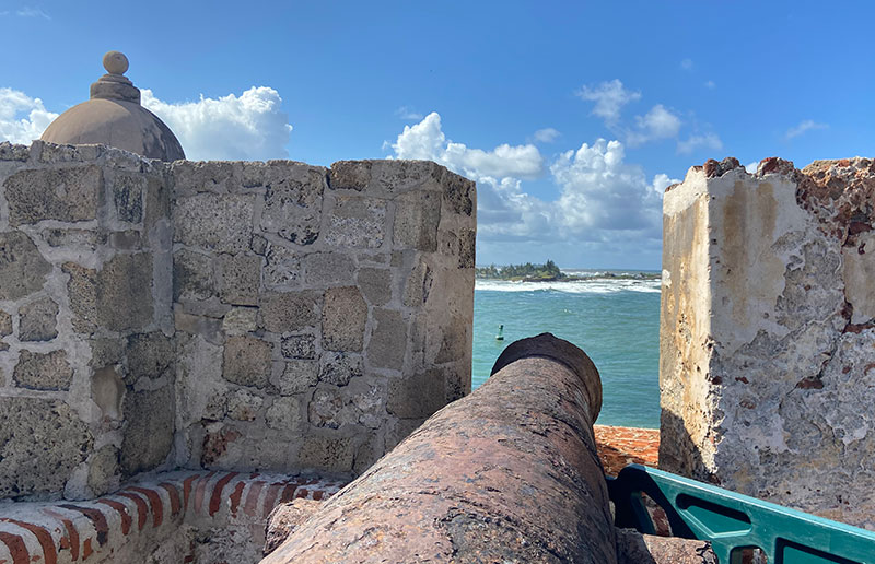 A rusted iron cannon pointing out through the stone battlements of a historic fort toward the ocean.