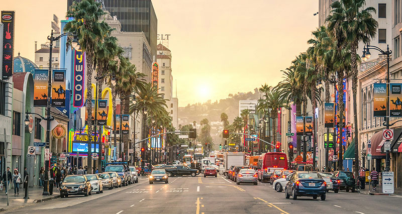 A busy street view of Hollywood Boulevard in Los Angeles at sunset, lined with tall palm trees, cars, and illuminated theater signs.