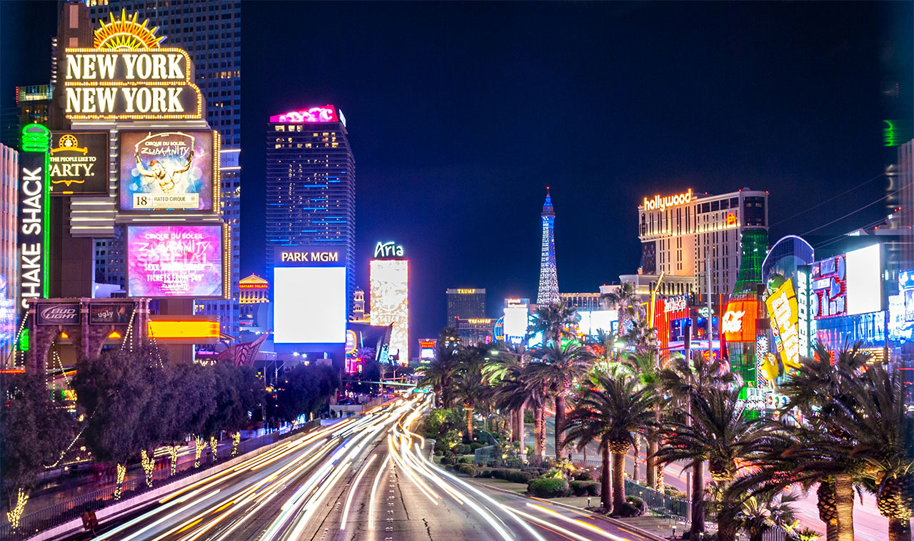 A vibrant night view of the Las Vegas Strip, featuring the illuminated Eiffel Tower at Paris Las Vegas, the High Roller observation wheel, and the glowing neon lights of the surrounding resorts.