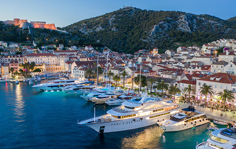 Aerial view of Hvar Town harbor at twilight, filled with luxury yachts and sailboats with an illuminated hillside fortress in the background.