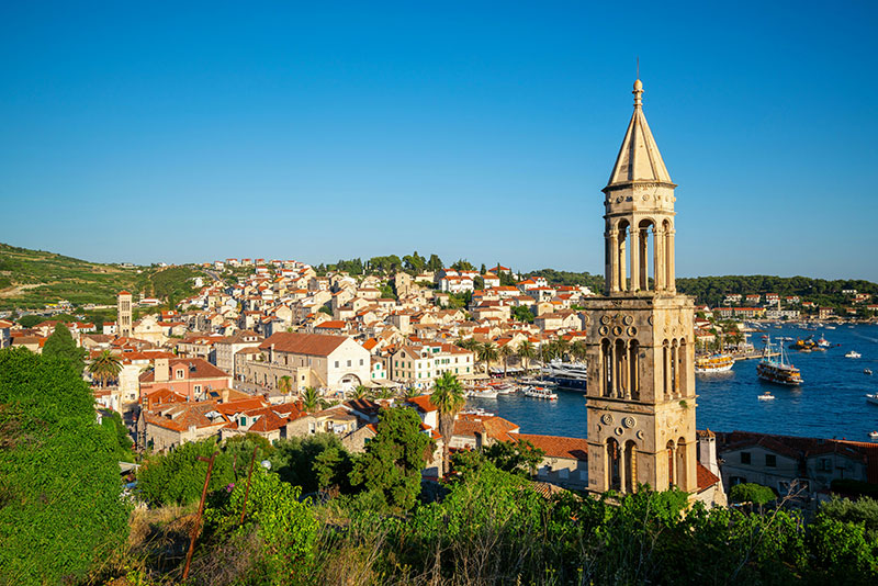 Elevated view of Hvar Town on a sunny day, featuring the stone bell tower of St. Stephen's Cathedral, terracotta roofs, and boats in the harbor.