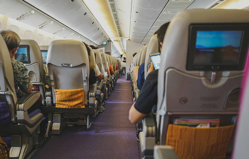 An interior view of an international flight cabin, showing rows of seating with passengers, seatback entertainment screens, and a long aisle stretching toward the back of the airplane.
