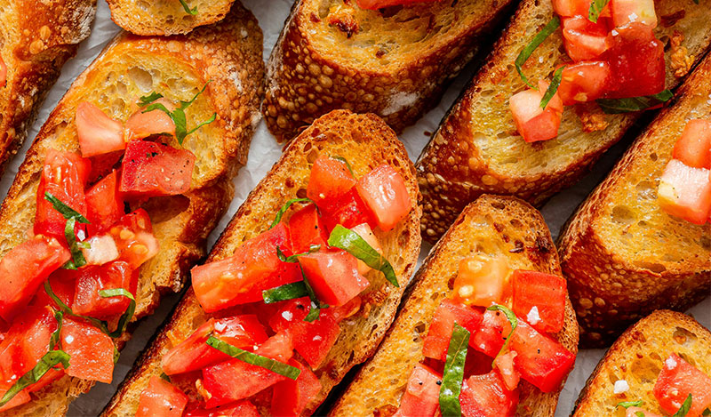 A close-up of several toasted bruschetta slices topped with fresh diced tomatoes and green herbs, served on a white plate during an Italian aperitivo