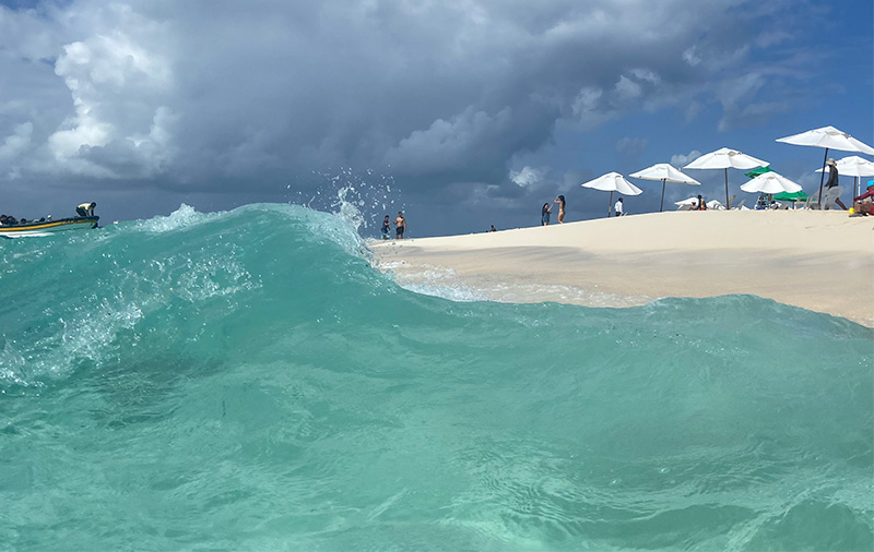 A bright turquoise wave crashing on a white sand beach with sun umbrellas in the background on Johnny Cay