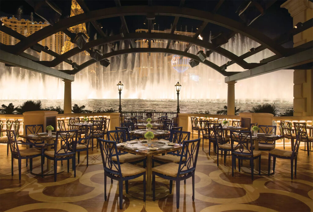 An elegant outdoor dining patio at night with a view of the Bellagio Fountains, featuring set tables under a dark wooden pergola with hanging lights