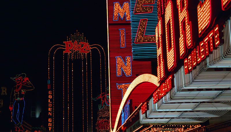 A close-up night view of vintage neon signs in old downtown Las Vegas, featuring glowing red and orange lights from classic casino fronts like the Mint and Horseshoe.