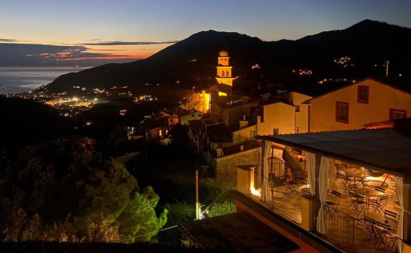 A hillside view of Levanto, Italy at dusk, featuring warm glowing lights from traditional buildings and a church bell tower against the silhouette of dark mountains under a deep blue twilight sky.