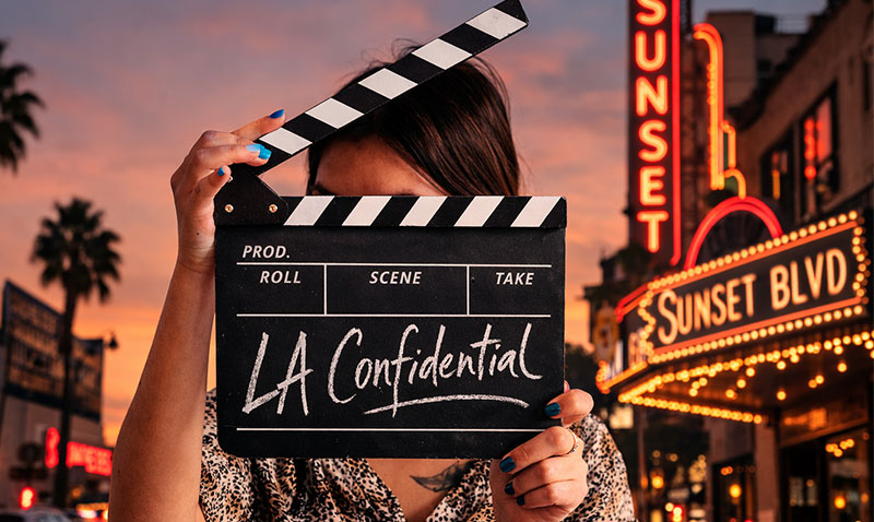 Film clapperboard reading “L.A. Confidential” held in front of Sunset Boulevard neon signs at dusk in Los Angeles