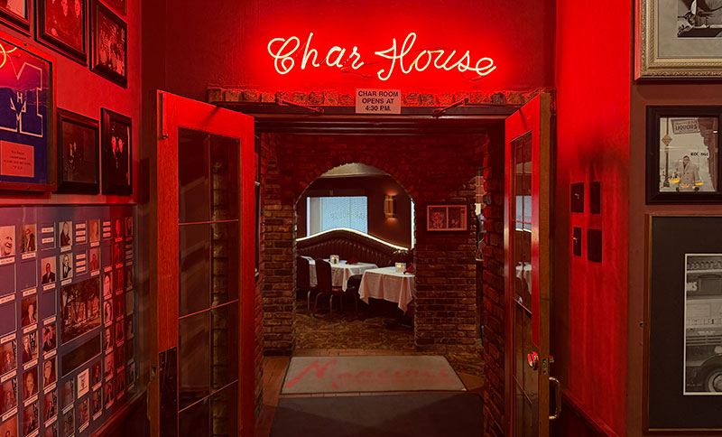 The glowing red neon Char House sign hanging above a brick archway leading into the main dining room at Mancini's.