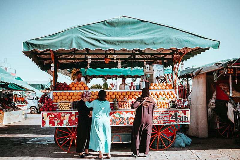 A vibrant street cart loaded with fresh oranges under a green awning, with local women ordering freshly squeezed juice.