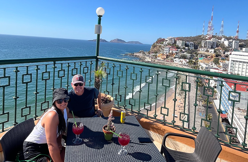 A couple sits at a sunny rooftop table with drinks, overlooking the coastal malecon and beaches of Mazatlán.