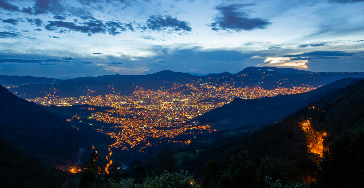 Nighttime aerial view of the Medellin city skyline glowing with warm lights in the valley, surrounded by the dark silhouettes of the Andes mountains at twilight.