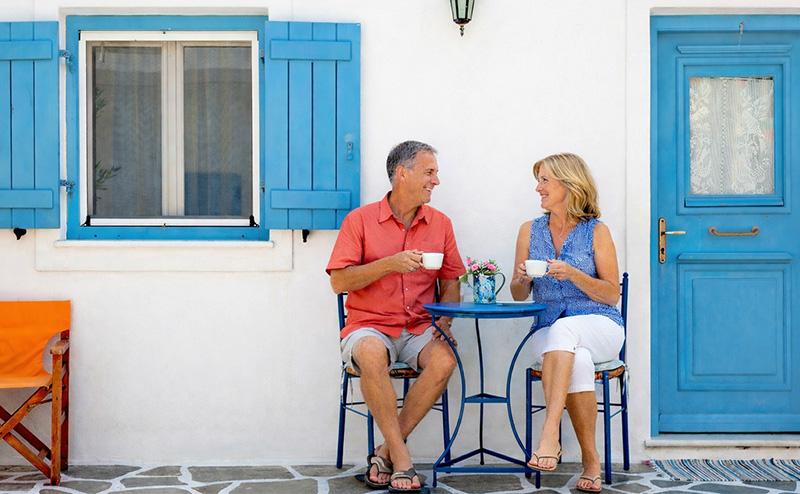 Retired expat couple sitting at a small table with coffee outside a white Mediterranean house with blue shutters and door in Greece.