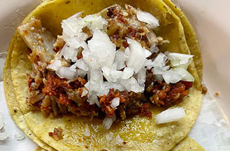 A close-up of two traditional Mexican street tacos on a plate, topped with fresh cilantro, onions, and white cheese, with a hand squeezing a fresh lime wedge over the